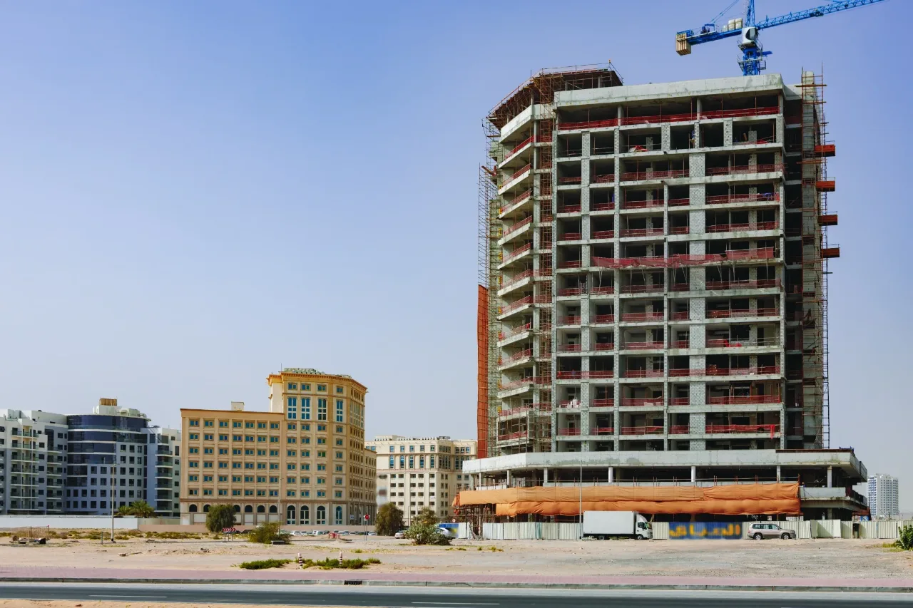 مبنى متوسط الارتفاع قيد الإنشاء على قطعة أرض مطلة على الطريق، تحيط به السقالات وسياج الموقع وتقف عند قاعدته شاحنة توريد