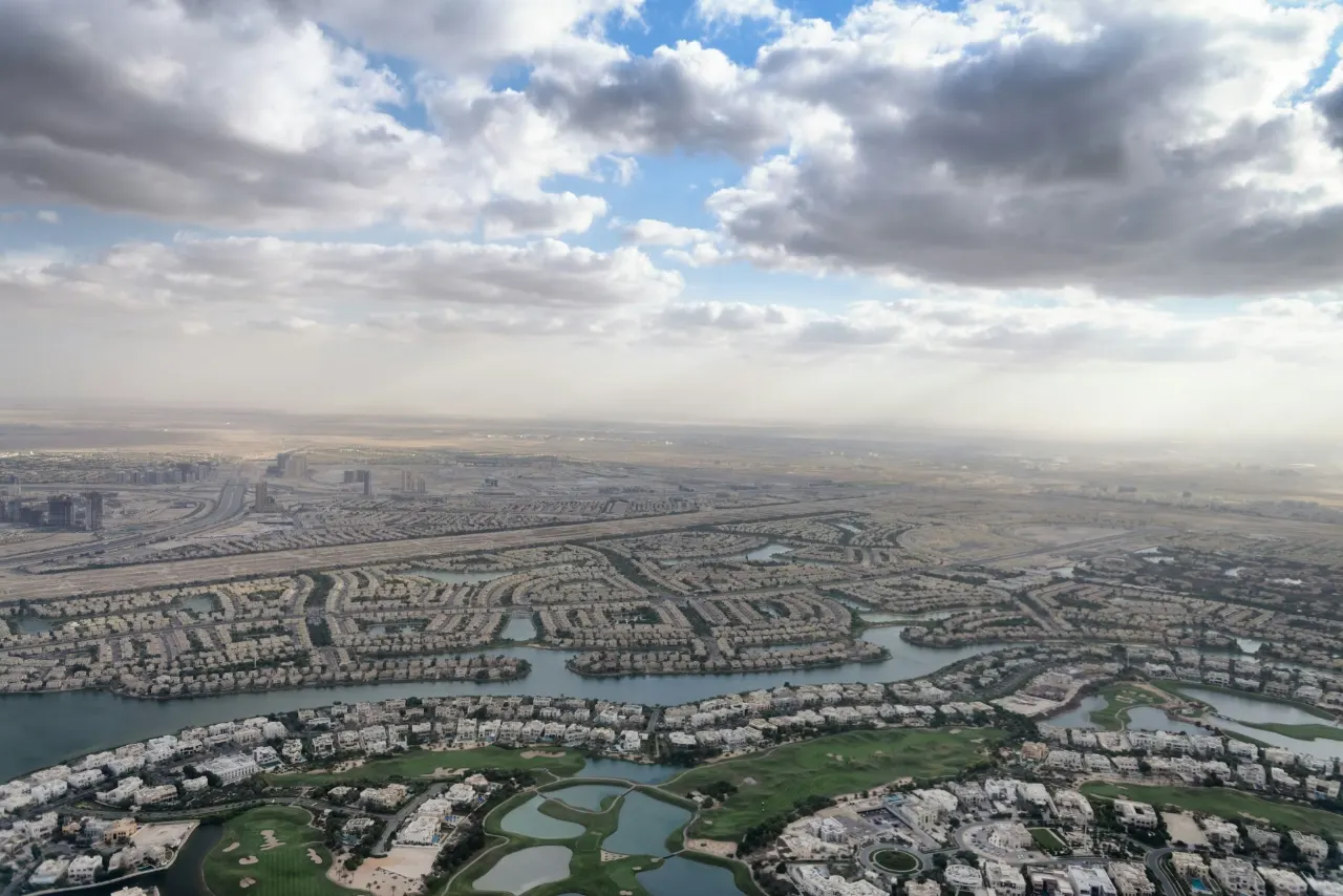 منظر جوي لأطراف دبي، حيث تلتقي الأحياء السكنية والممرات المائية ومحاور الطرق بالصحراء المفتوحة تحت سماء غائمة جزئياً